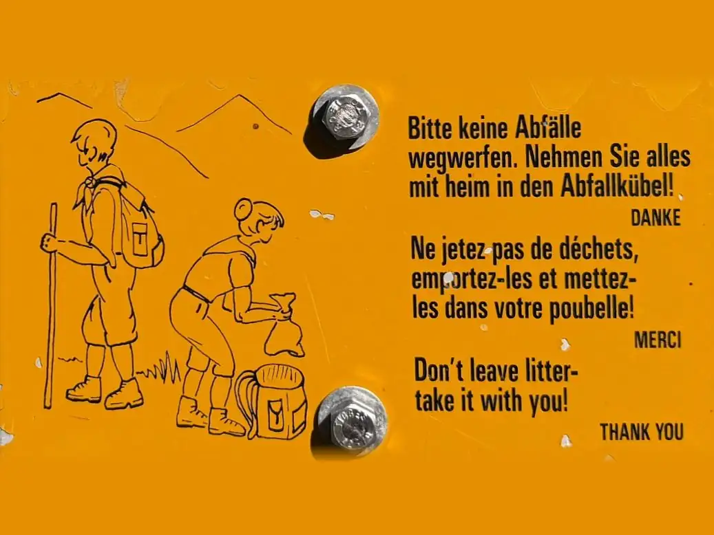 Gelbe Hinweistafel am Wanderweg mit zwei gezeichneten Wandernden: «Bitte keine Abfälle wegwerfen. Nehmen Sie alles mit heim in den Abfallkübel!», dazu auf Französisch und Englisch